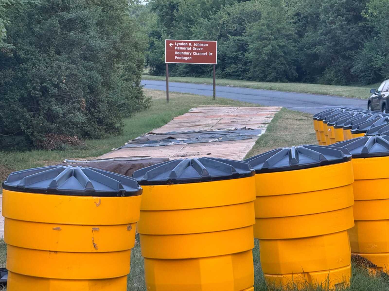 Row of bright yellow waste bins with gray lids lined up along a grassy path by a road and green trees, with a red sign in the background.