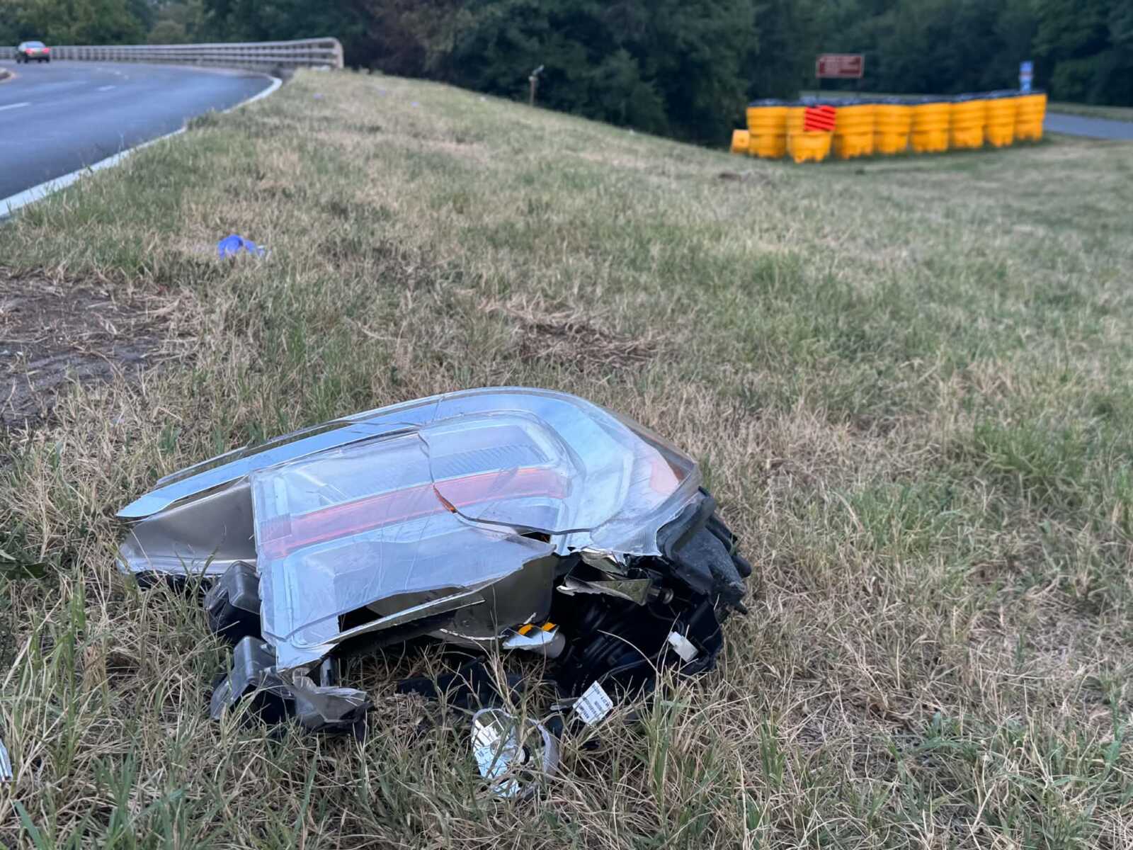 Crushed silver car wrecked in a grassy median beside a curving highway, debris scattered nearby.
