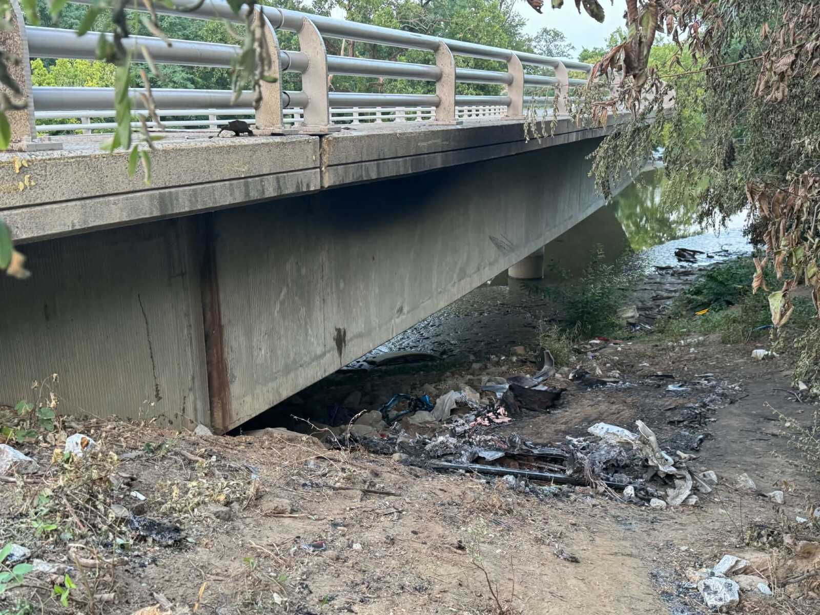 Bridge over a polluted canal with a litter-strewn bank beneath and debris scattered on the ground nearby, undergrowth framing the scene