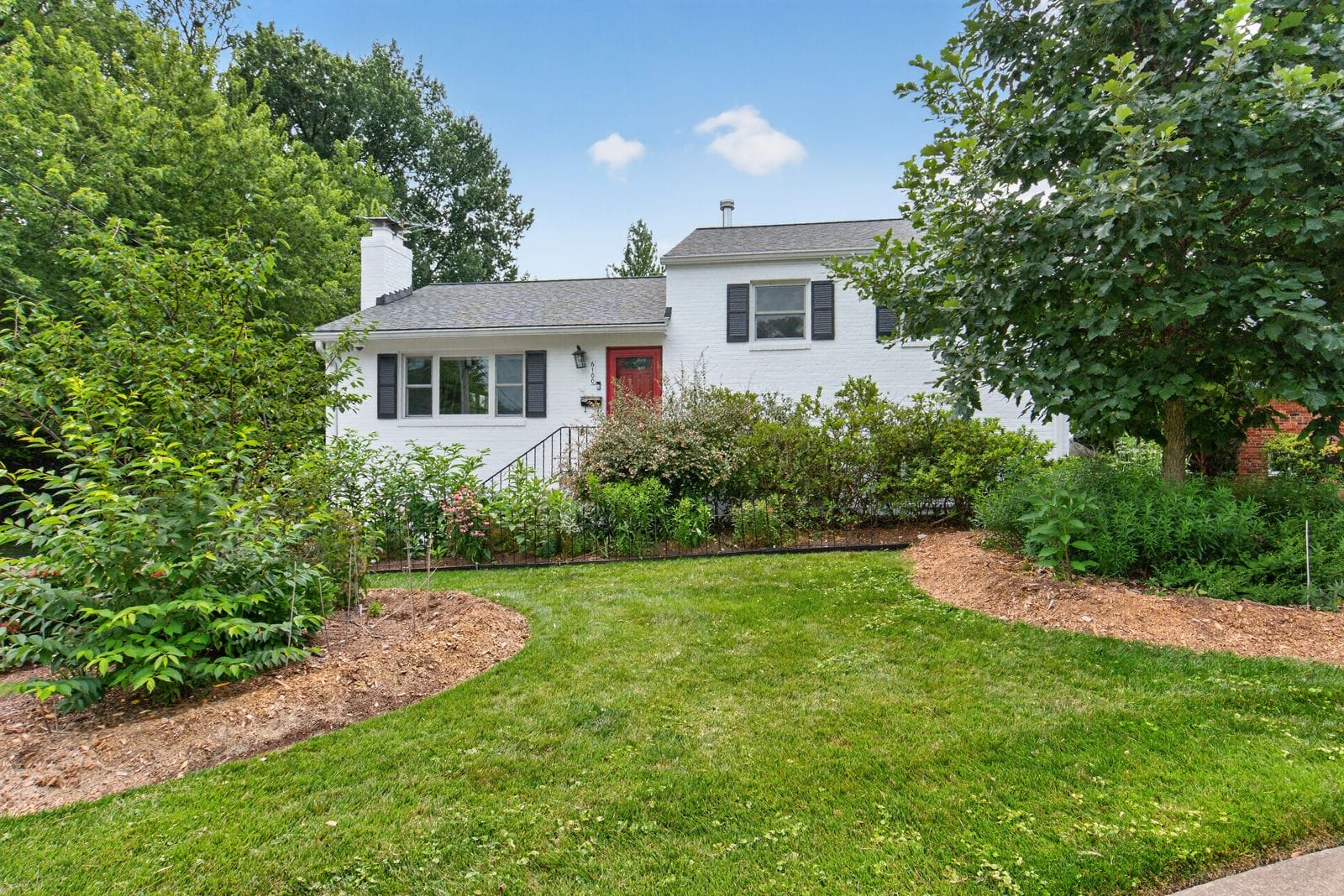 White brick house with red front door, black shutters, and a manicured garden in a sunny yard.