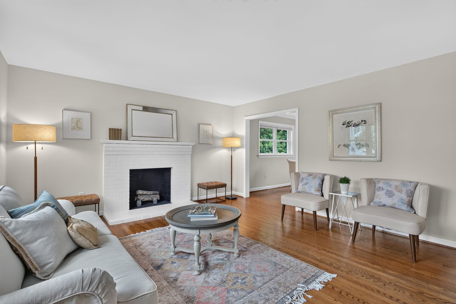 Bright living room with a white brick fireplace, gray walls, and warm wood floors. A round coffee table sits on a patterned rug beside a sofa and chairs.
