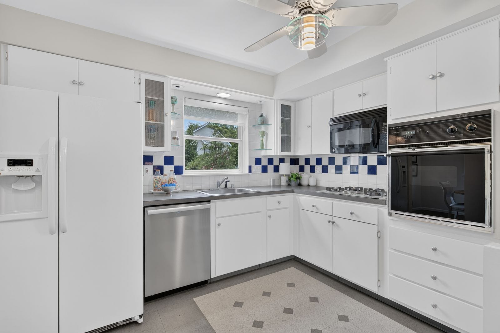 Bright white kitchen with white cabinets, stainless dishwasher, and a blue-and-white checker tile backsplash under a window above the sink.