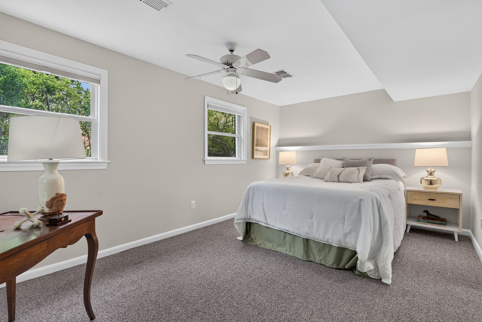 A calm bedroom with a white bed, beige walls, two nightstands, and a ceiling fan, natural light from two windows.