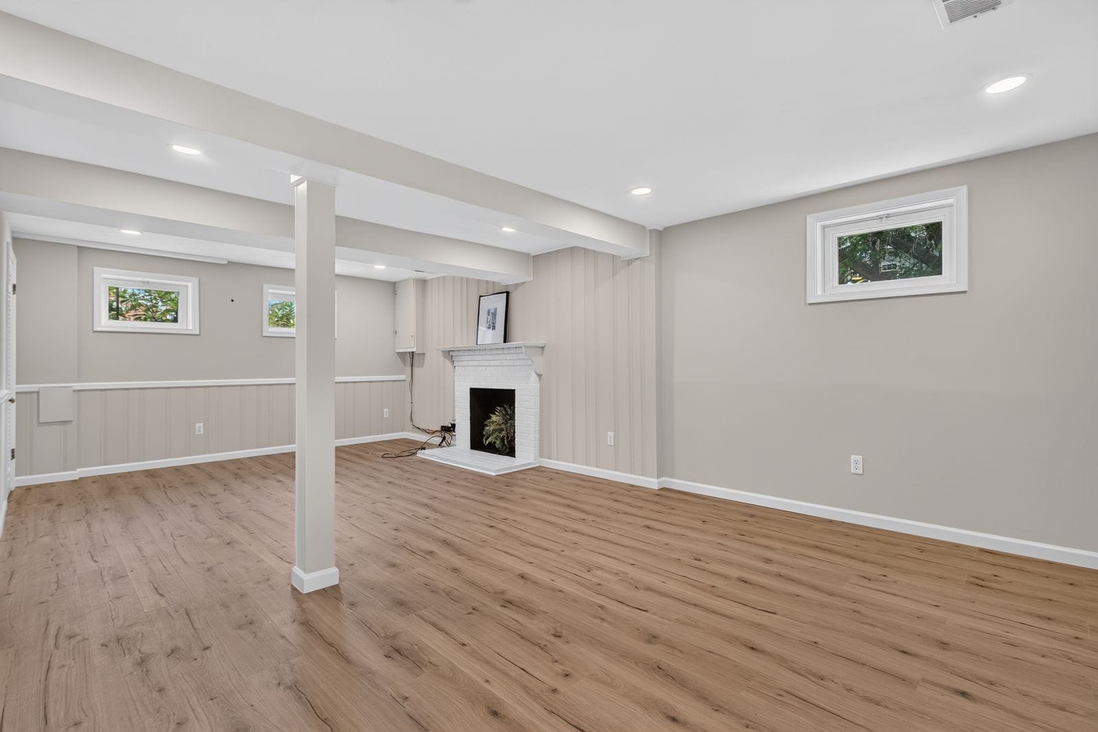 Bright finished basement with white beams, beige walls, and a white brick fireplace. Hardwood-look floor.