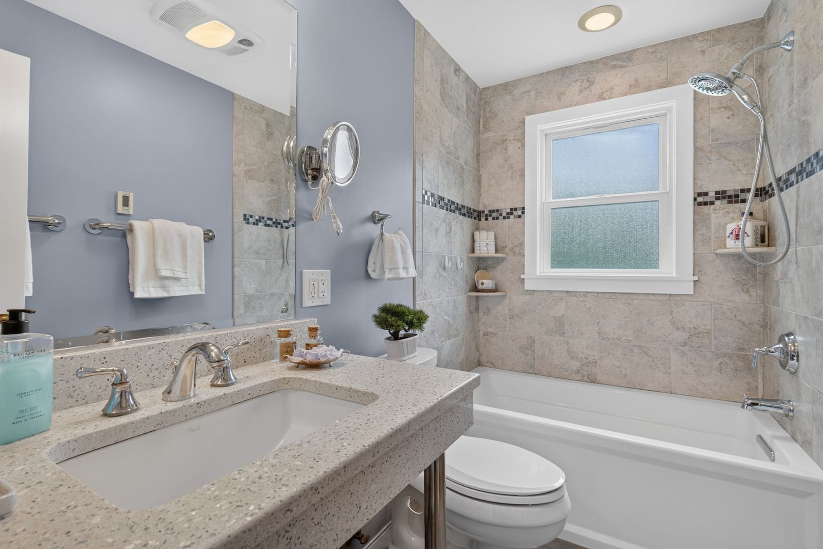 Bright modern bathroom with blue walls, speckled quartz countertop, and a tiled shower/bath with a frosted window and chrome fixtures up front.