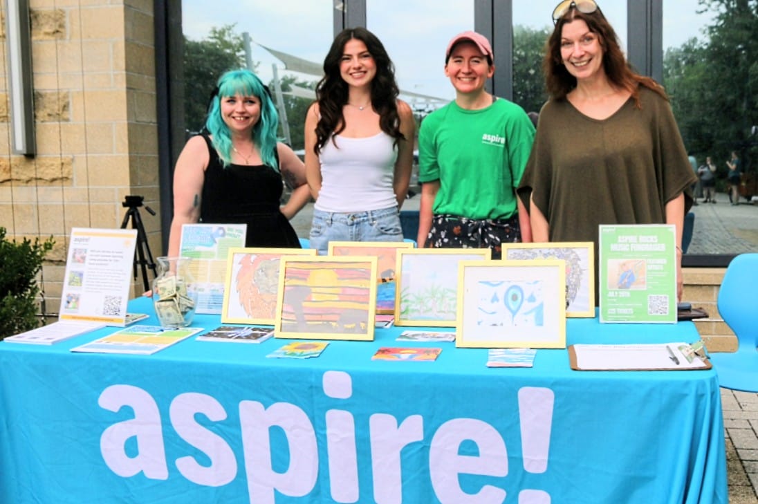 Four women stand behind a bright blue table with framed artwork and brochures at an outdoor event, smiling at the camera.