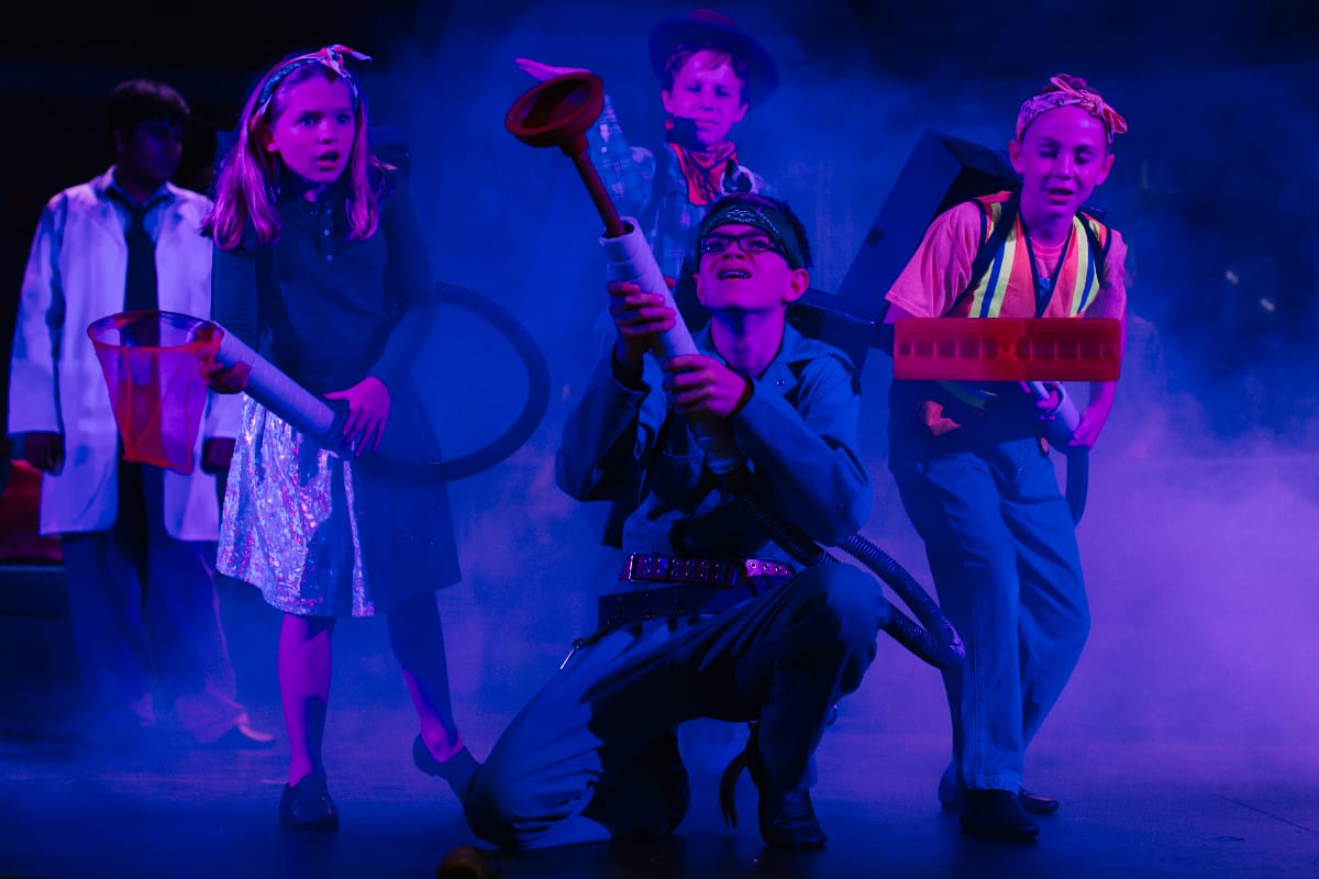 Children in a stage play, dressed in costumes, holding props under blue stage lighting with fog.