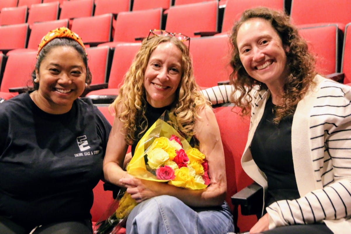 Three women smiling in a theater seats area; the center woman holds a bouquet of yellow and pink roses wrapped in yellow paper inside red seats of a venue.