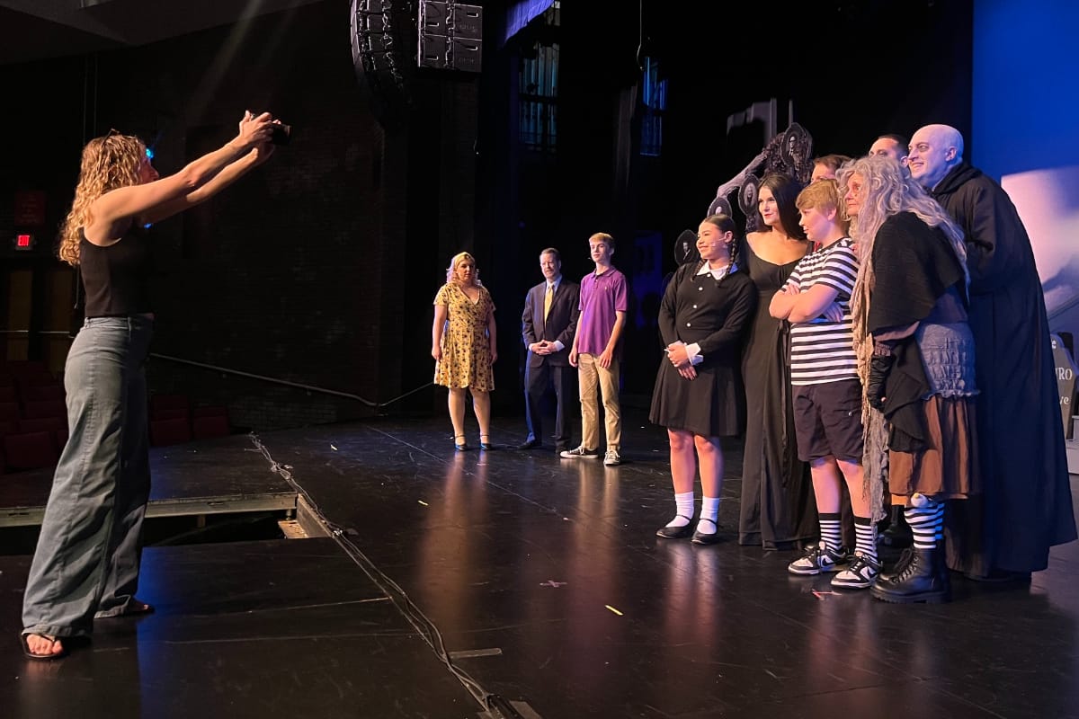 Woman on stage taking a photo of a line of performers in costumes during a show on a theater stage.