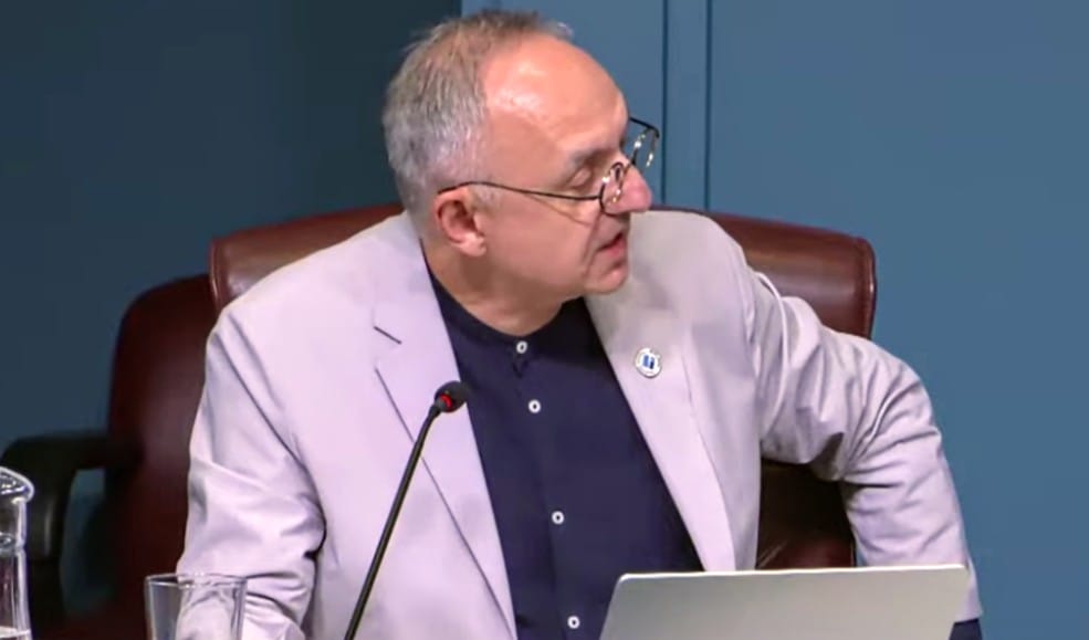 County Board member Takis Karantonis responds to Susan Cunningham at July 21, 2026, meeting (screenshot via Arlington County) | ARLnow Man in a light blazer and dark shirt sits at a desk with a microphone and laptop during a meeting or panel.