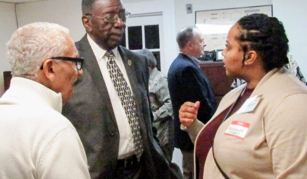 Professional networking scene: two men and a woman in business attire converse in a hallway, the woman wearing a name badge and beige blazer.