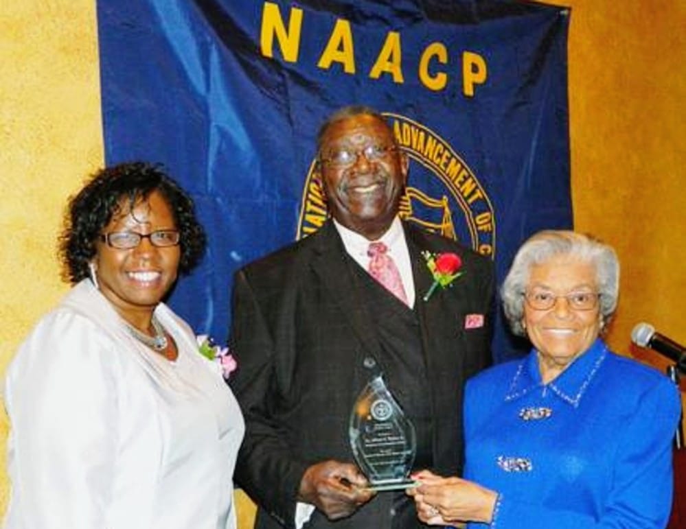 Three people at an awards ceremony posing with a glass trophy in front of a NAACP banner, the man center wearing a suit and pink tie as the award recipient.
