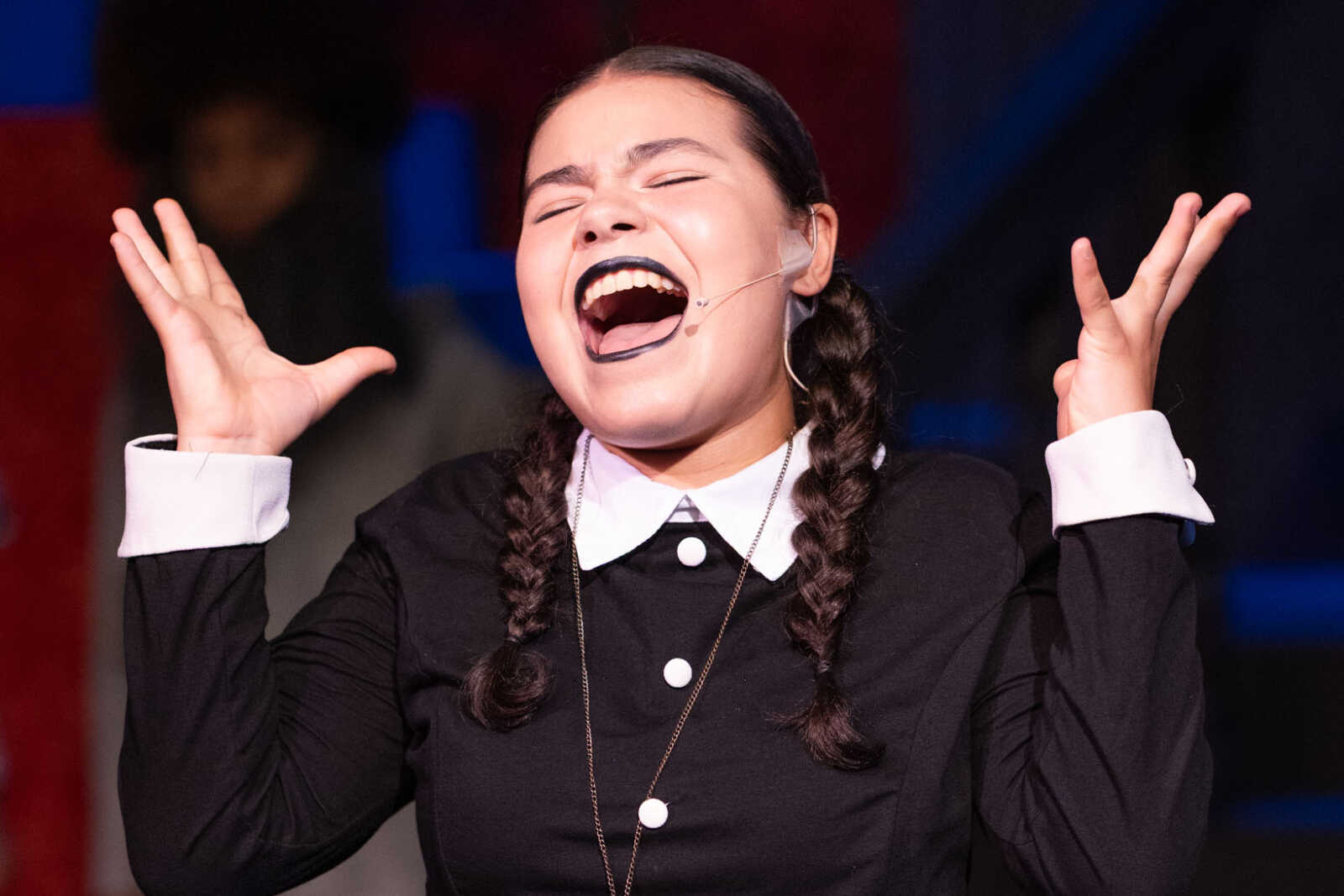 Performer on stage laughing with mouth wide open, wearing a black outfit with white collar and braided hair, headset mic visible