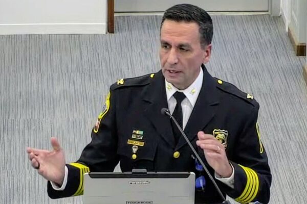 Uniformed officer speaks at a podium during a briefing, gesturing with his hands while addressing an audience.