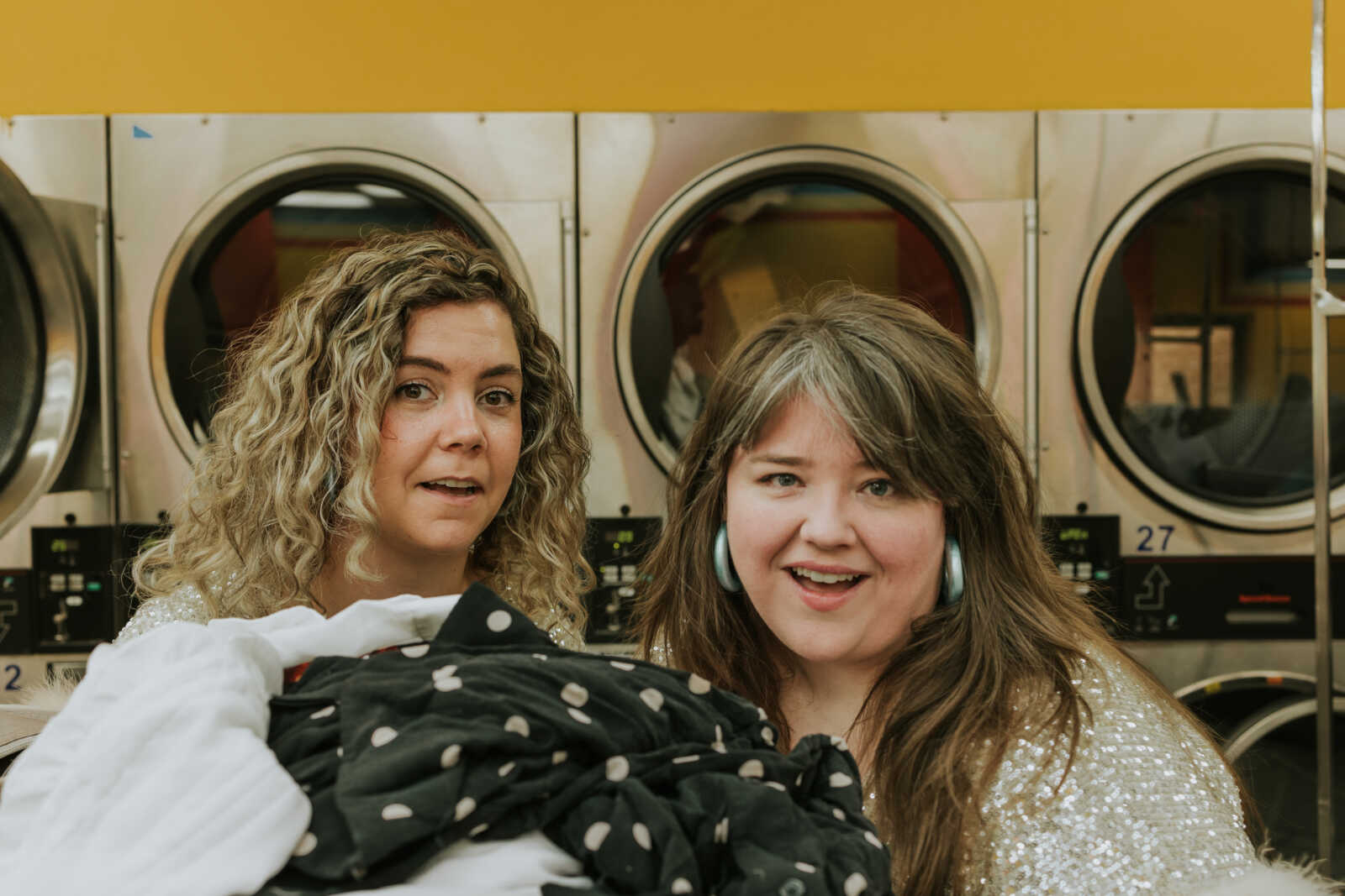 Two women smiling in a laundromat, holding a pile of laundry with front-loading machines behind them on a yellow wall.