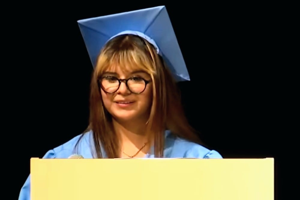 Helen Morataya Centeno speaks at the summertime graduation ceremony (courtesy APS) | ARLnow Graduate in a blue cap and gown giving a speech behind a podium, smiling at the audience.