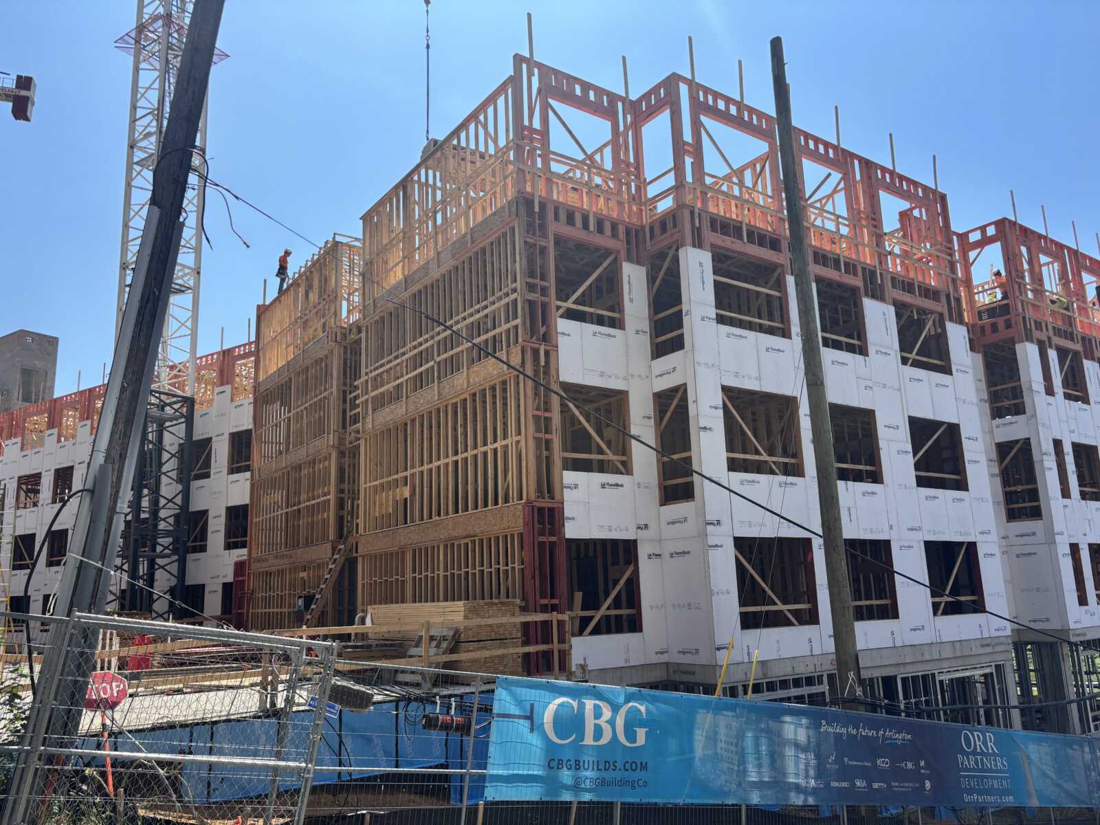 Active construction site; a multi-story wooden-frame building being erected under a clear blue sky, with a crane and scaffolding visible.