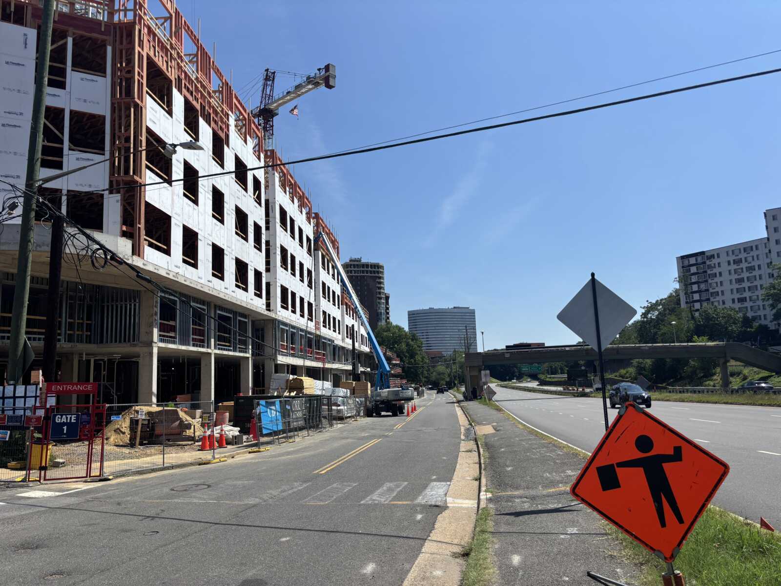 Construction site with a tall building under construction and a crane on the left; road in the foreground with safety barriers and signs.