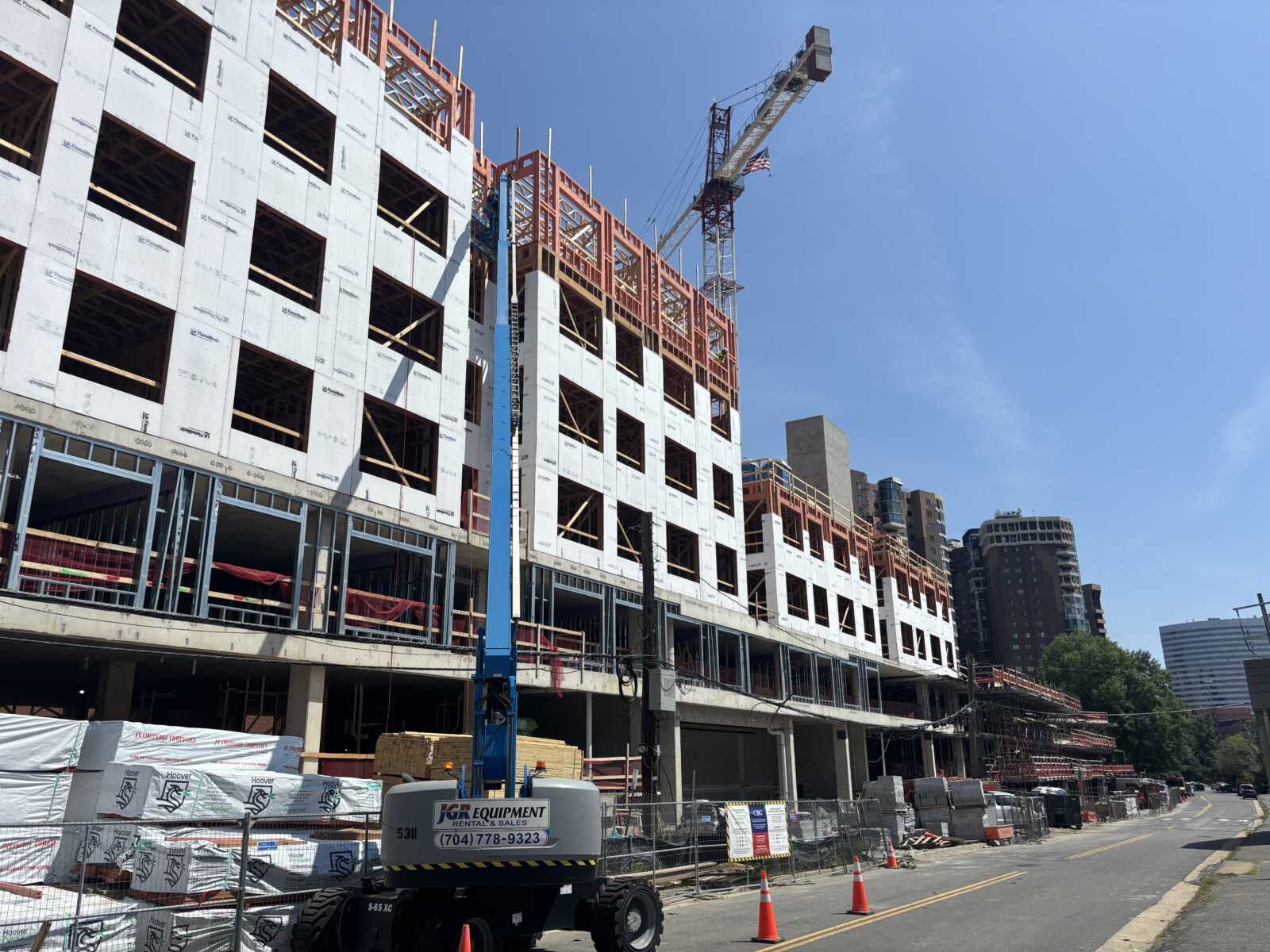 Construction site with a multi-story building under construction, white sheathing and orange framing, crane tall in the blue sky above