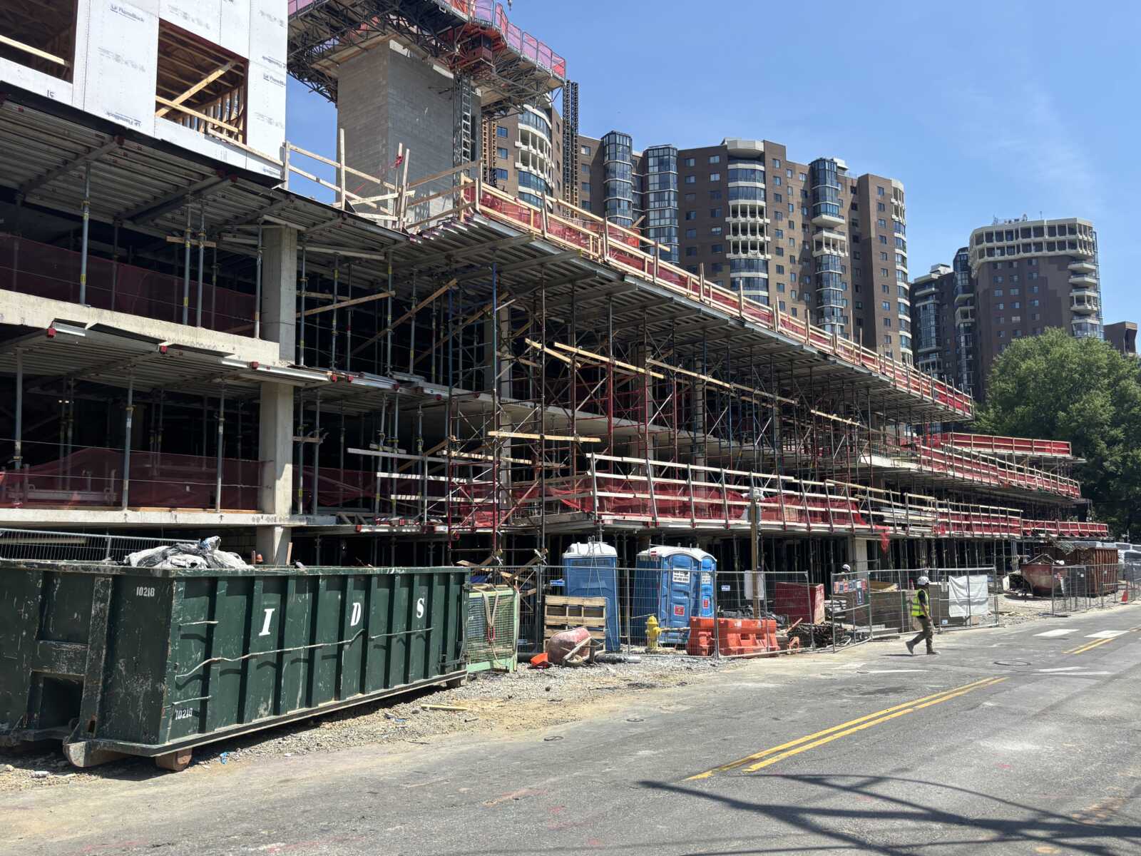 Construction site with scaffolding along a multi-level building under construction; red safety rails, portable toilets, and a large dumpster in the foreground with city high-rises in the background.