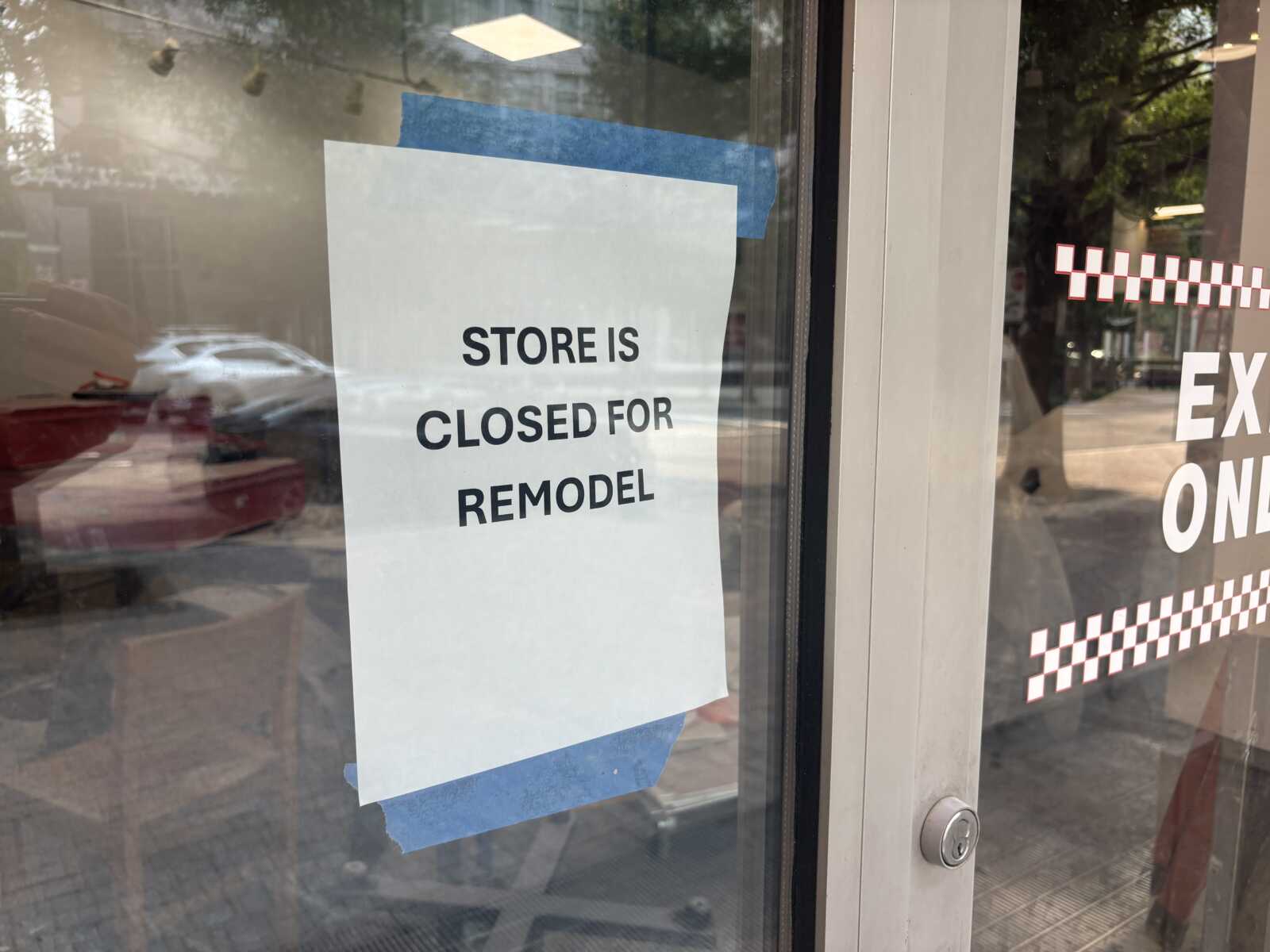 White paper sign on a glass storefront reads 'STORE IS CLOSED FOR REMODEL' taped with blue painter's tape, with street reflections visible behind.