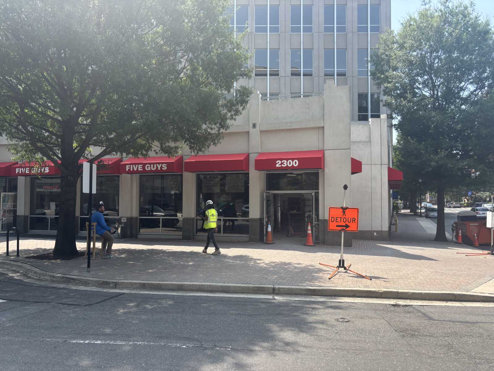 Five Guys restaurant with red awnings on a city street, detour sign and construction cones nearby amid pedestrians and trees.