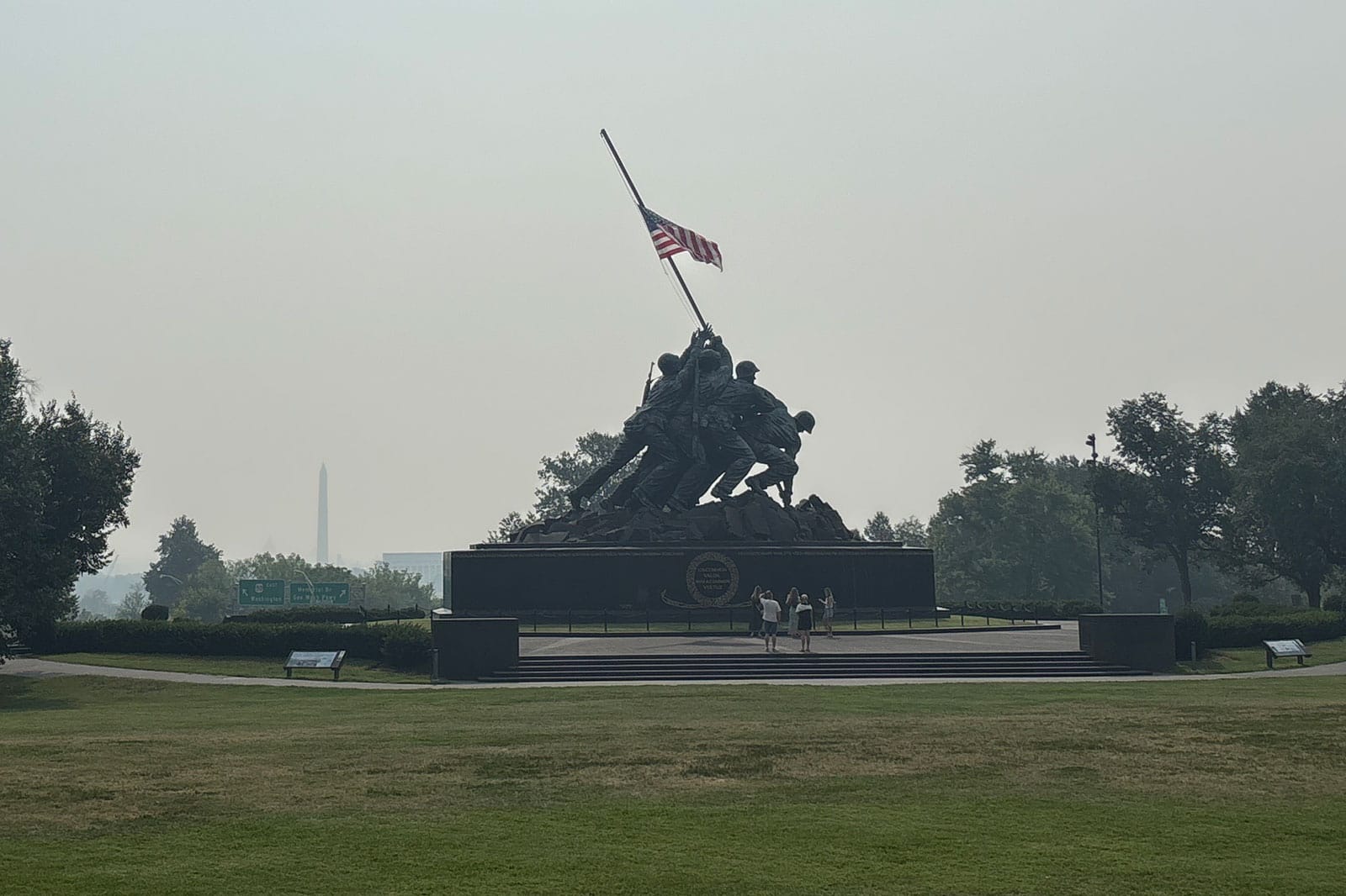 Silhouetted soldiers raise a flag atop a rocky monument; distant Washington Monument visible on the horizon; visitors stand at the base on a lawn.