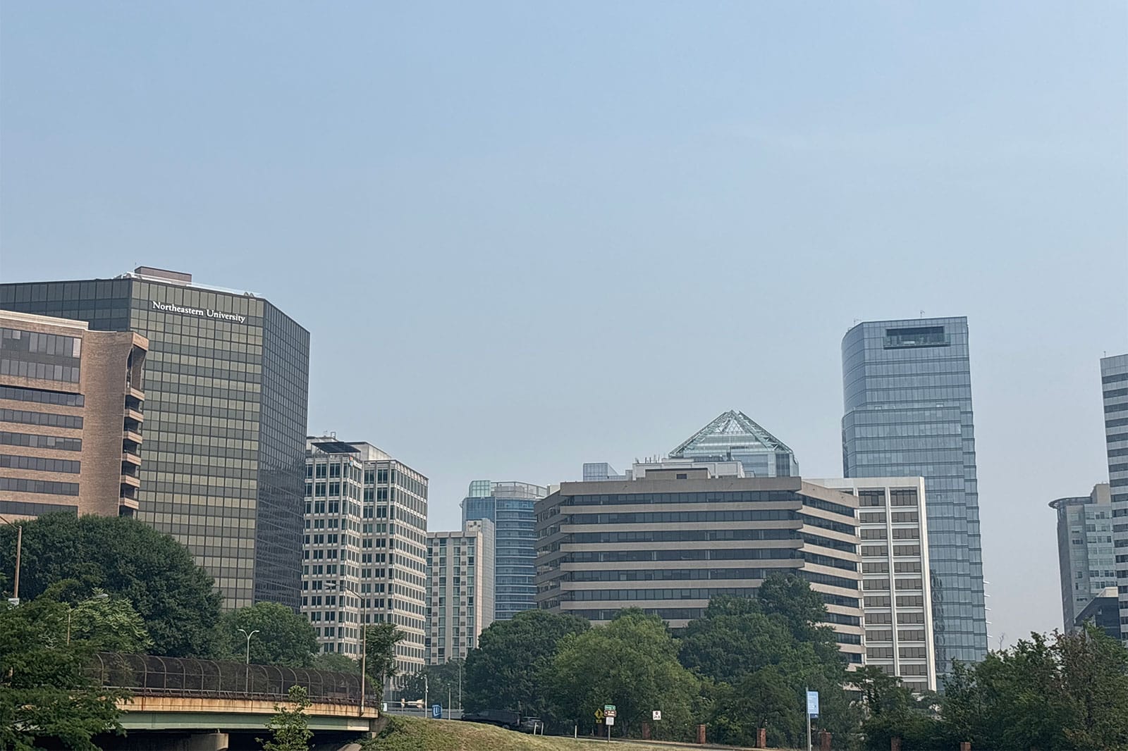 Buildings in Rosslyn amid wildfire haze (staff photo by Ryan Cole) | ARLnow City skyline with modern glass office buildings and clear blue sky; Northeastern University sign on the left building.