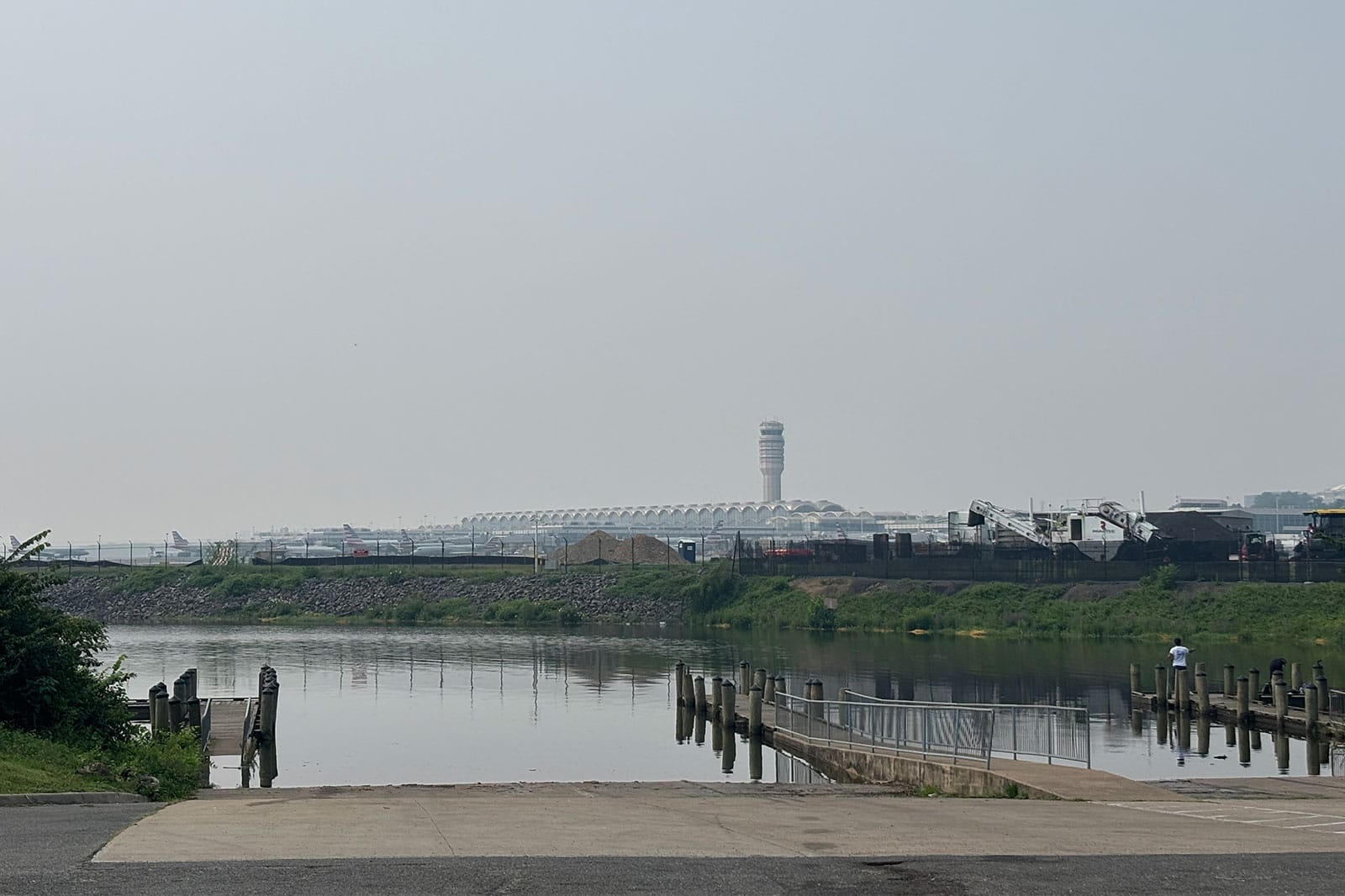 Reagan National Airport as seen from Gravelly Point, amid wildfire haze (staff photo by Ryan Cole) | ARLnow Industrial shoreline with a distant airport control tower, water in the foreground and construction activity along the bank.
