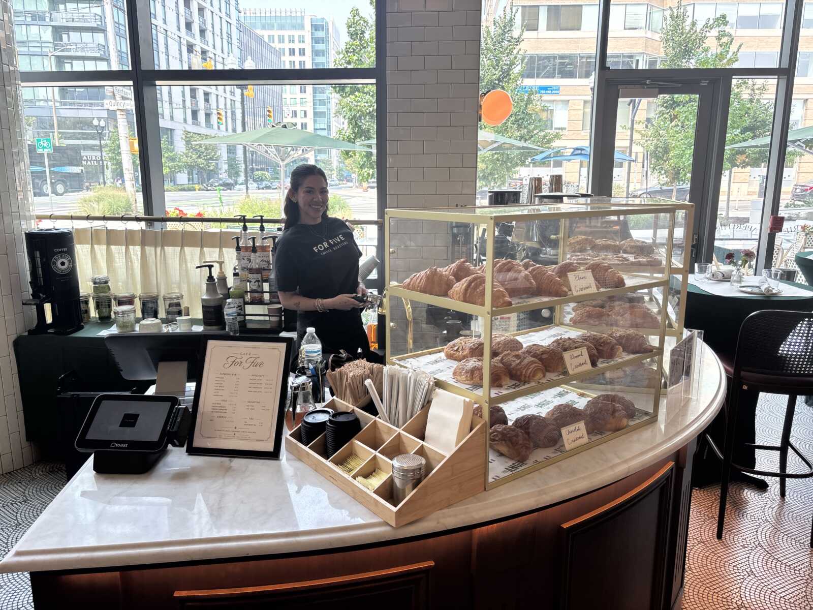 Smiling bakery staff behind a curved marble counter with a glass display case full of croissants and pastries in a sunny cafe window setting outside city streets.