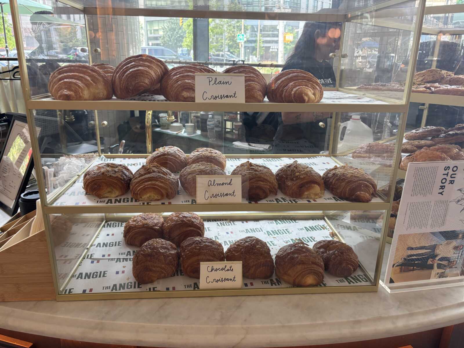 Display case of croissants labeled plain, almond, and chocolate croissants on two shelves in a bakery storefront.