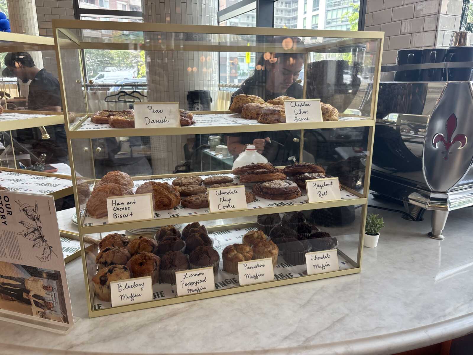 Glass bakery case with two shelves of assorted pastries and handwritten labels like Pear Danish and Cheddar Cheese Scone.