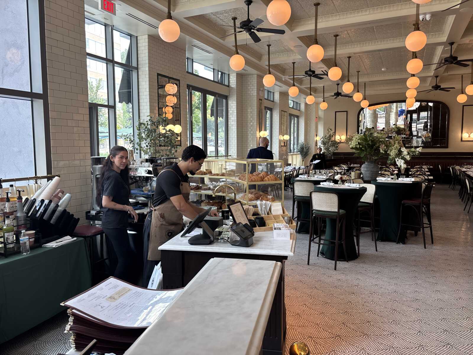 Bright cafe interior with a pastry display, two baristas at the front counter, and round pendant lights opening to large windows nearby.