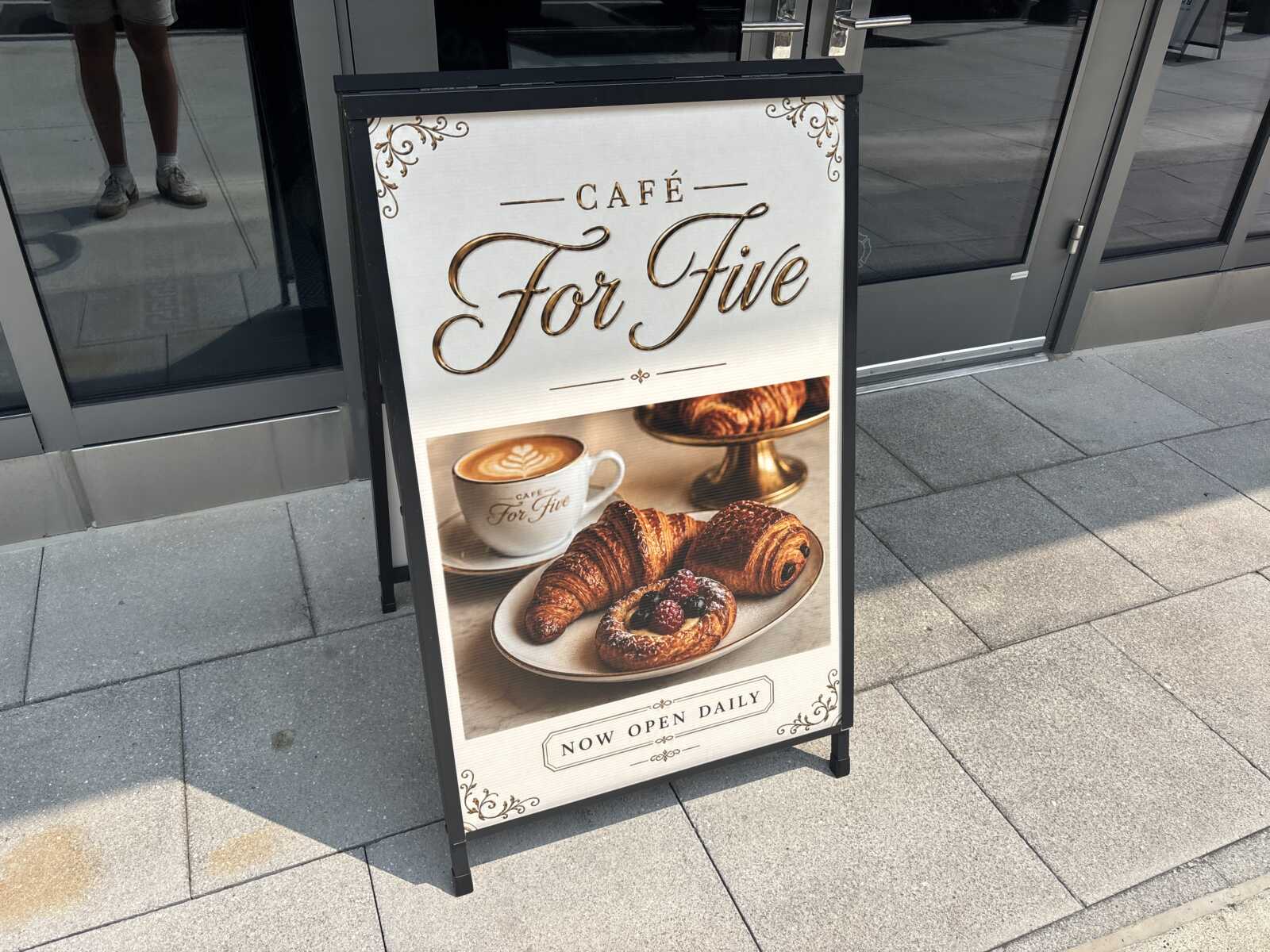 Outdoor sidewalk signboard advertising Café For Five with a cup of coffee and pastries (croissants) on a plate outside a cafe.