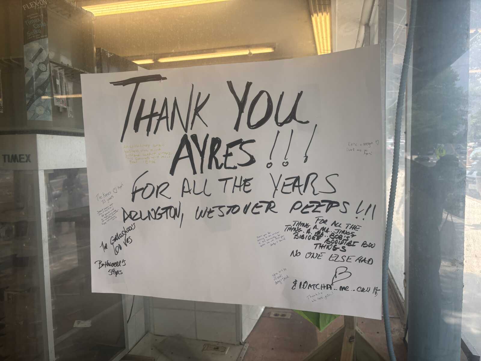 Large white poster in a storefront window with bold black handwriting that says 'THANK YOU AYRES for all the years' and small handwritten notes around the edges.