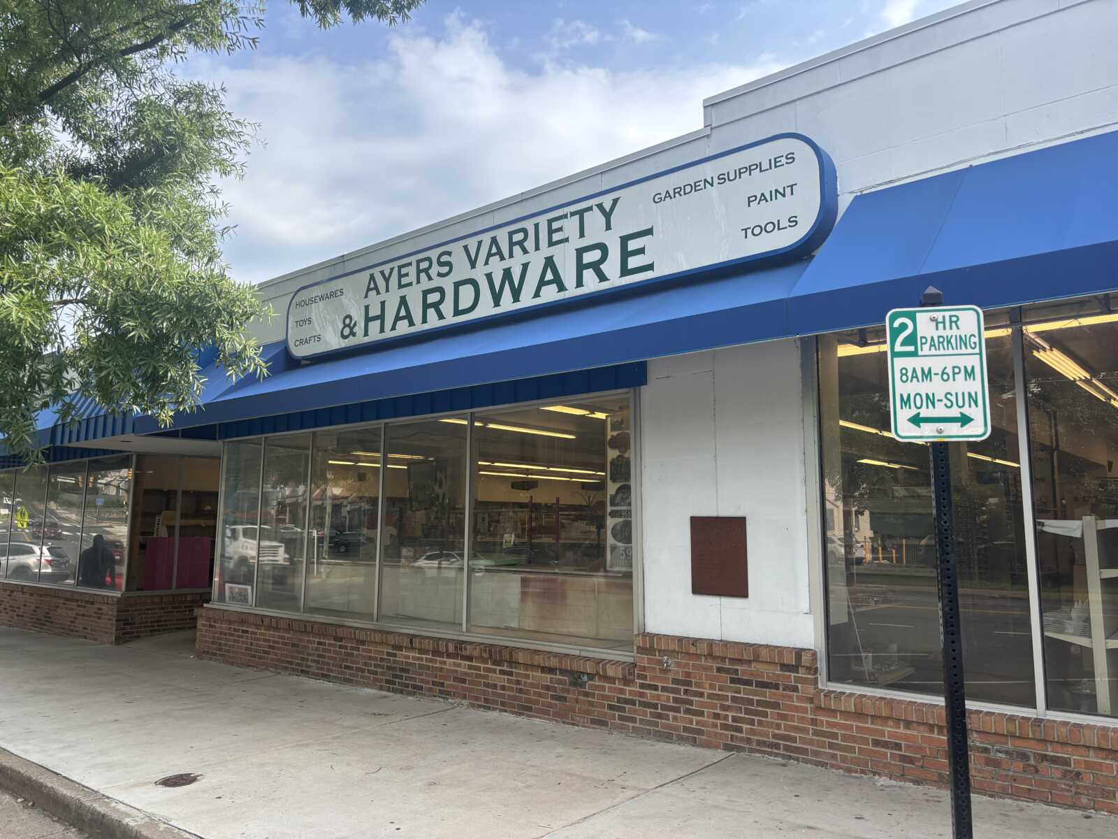 Storefront of Ayers Variety & Hardware with blue awning and a large sign, and a two-hour parking sign nearby.