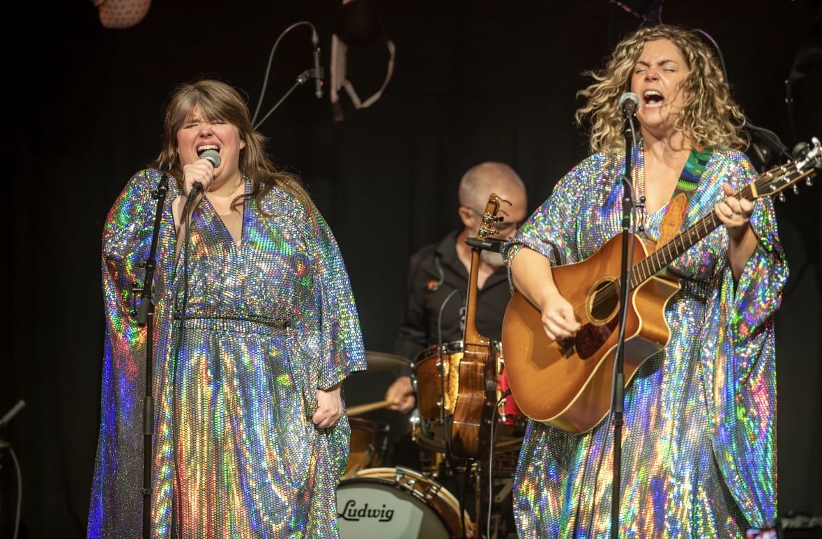 Two female singers in iridescent gowns perform on a stage with microphones, a guitarist to the right and a drummer in the background.