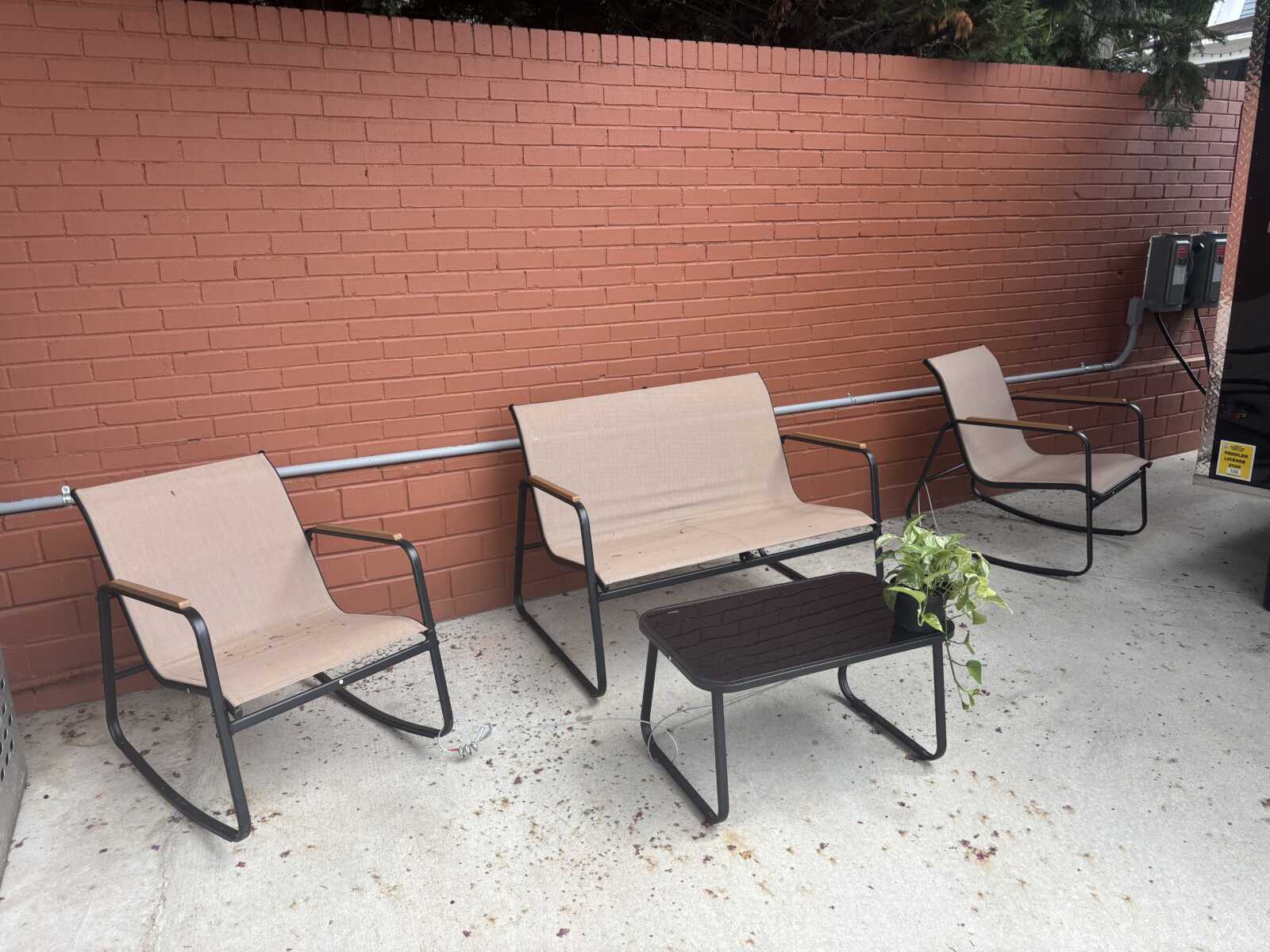 Outdoor patio with three beige mesh chairs and a small black coffee table tucked against a red brick wall, plus a potted plant on the table.
