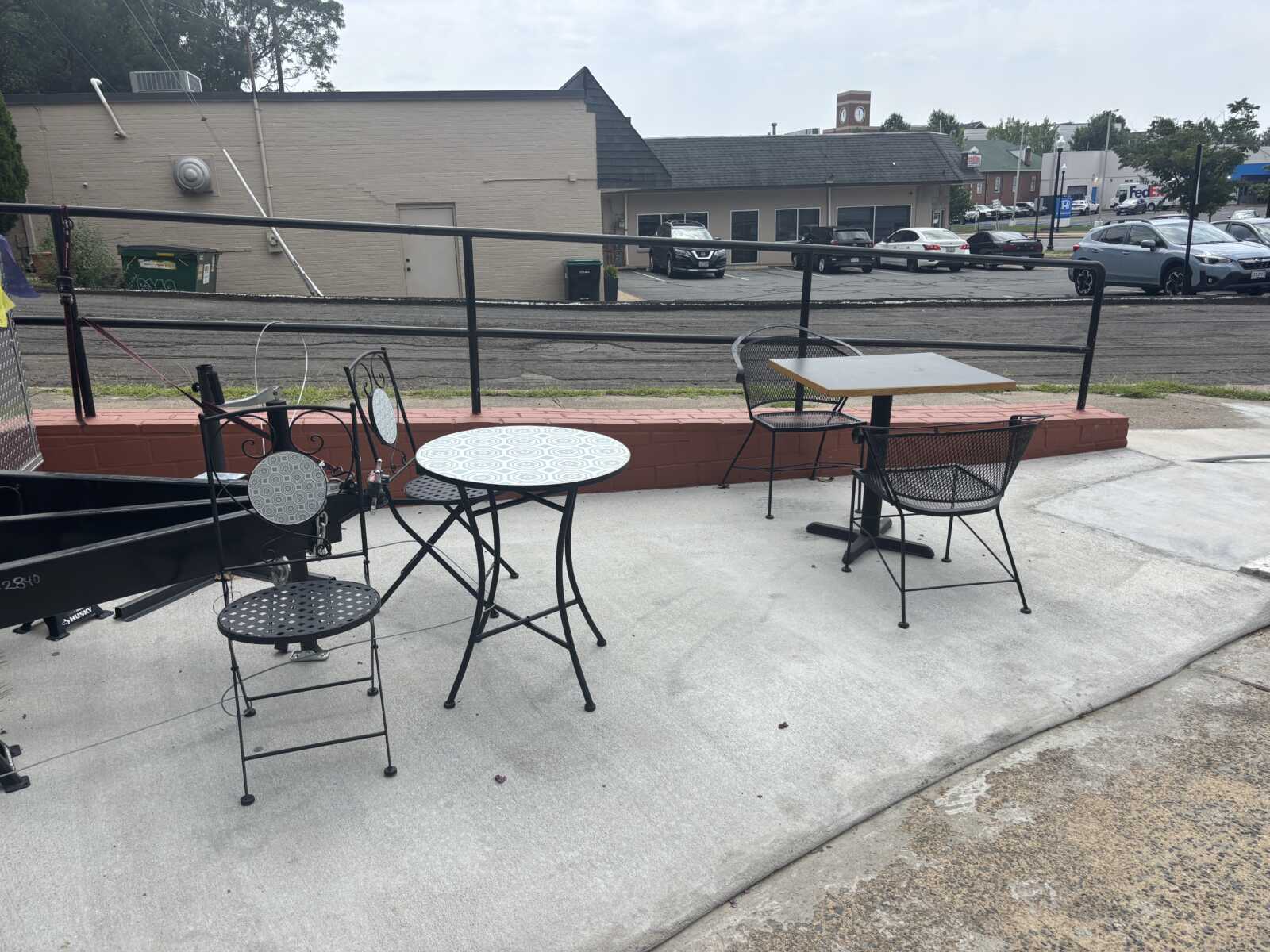 Outdoor seating area with black metal tables and chairs on a concrete patio next to a railing, overlooking a parking lot and street beyond.
