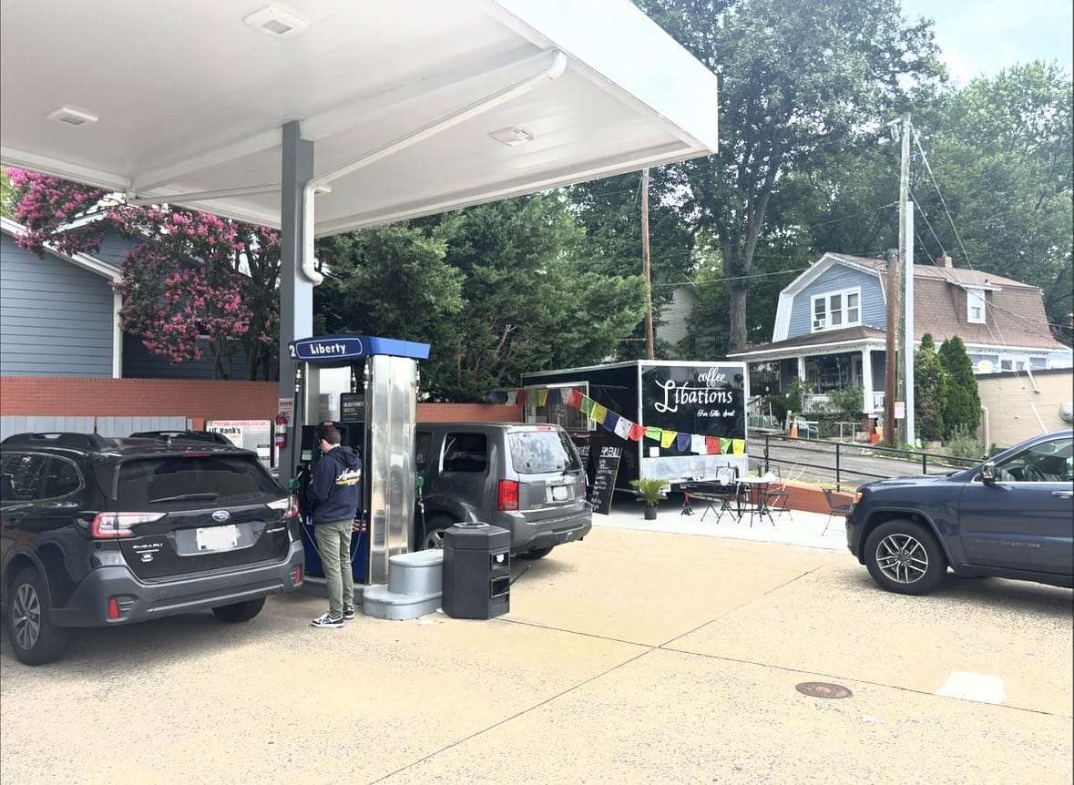 Gas station forecourt with a car refueling while a man stands at the pump; a coffee truck and outdoor seating nearby.