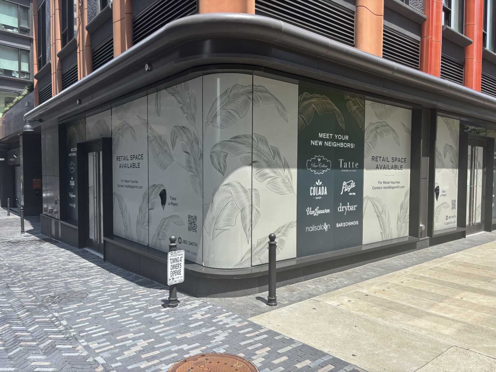 Corner storefront with glass panels covered in light gray tropical leaf graphics and ads for 'Retail Space Available' and 'Meet Your New Neighbors'.
