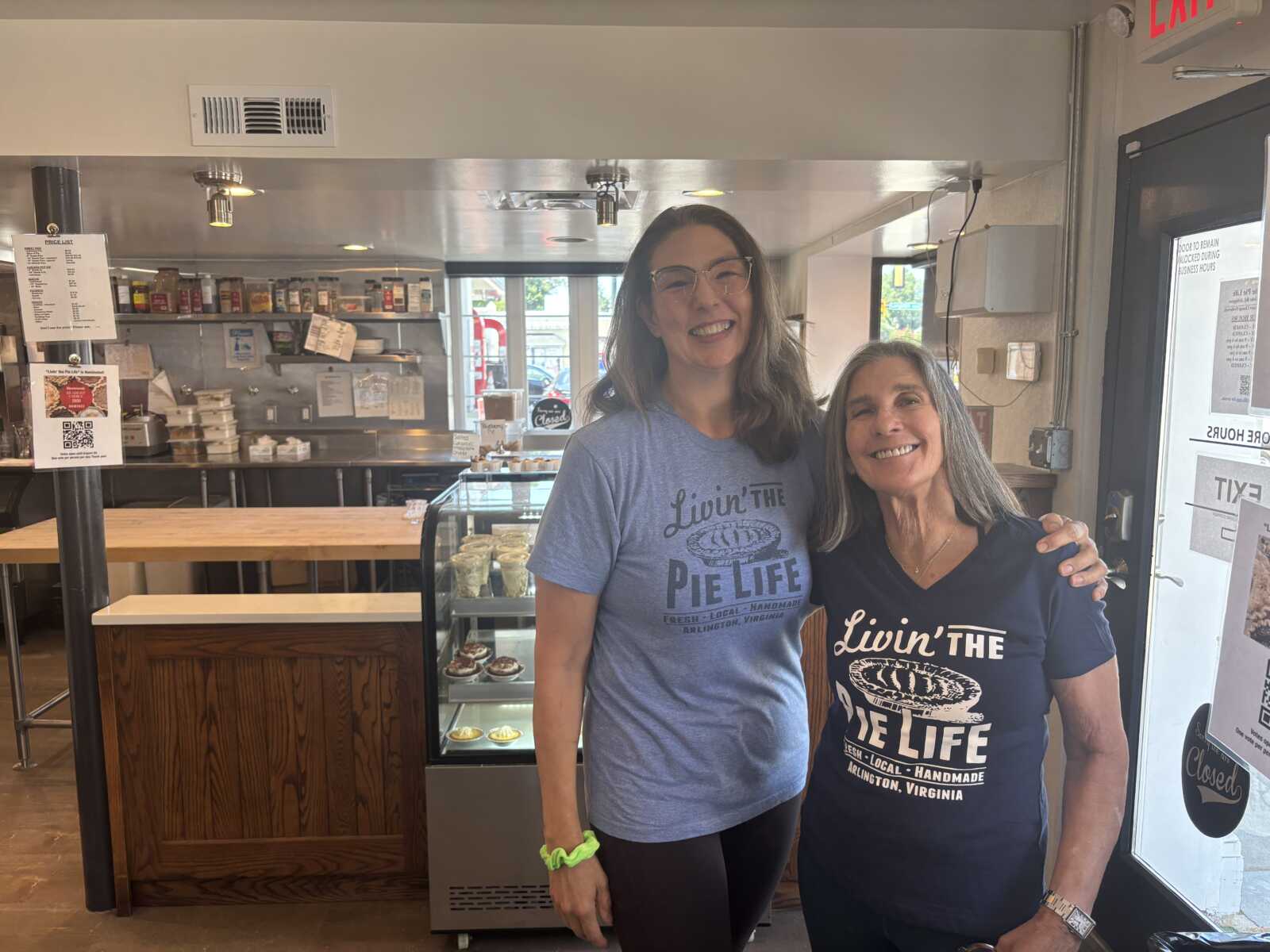Two women stand side by side in a bakery, smiling at the camera; one in a blue shirt and glasses, the other in a navy shirt that says 'Livin' the Pie Life'.