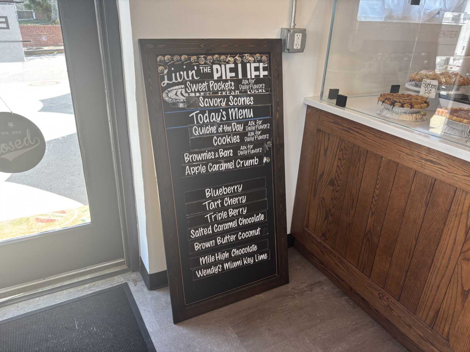 Chalkboard bakery menu outside the door listing pie and dessert flavors, like Blueberry and Apple Caramel Crumb.