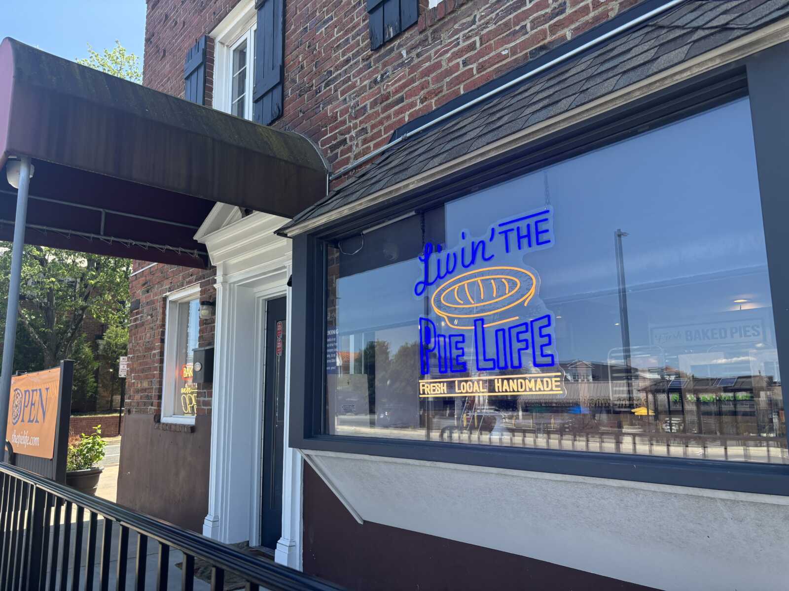 Brick storefront with neon sign reading "Livin' The Pie Life" in the window of a bakery; open sign visible outside.