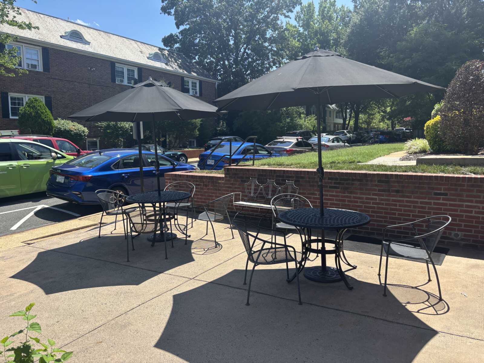 Outdoor cafe patio with round metal tables and chairs under large gray umbrellas, brick building and parked cars in background, sunny day.