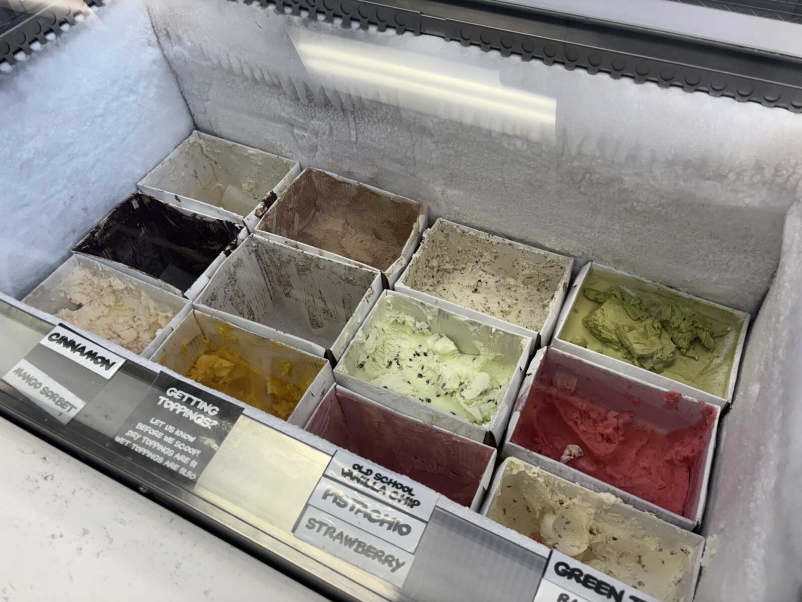 Palette of gelato flavors in metal tubs inside a display freezer, with pastel and vibrant colors like pink, yellow, green, and brown.