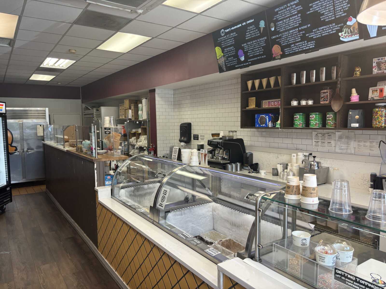 Interior of a cozy ice cream shop with a curved glass display case and a toppings shelf on the wall above the counter coasted by a white tile backsplash.