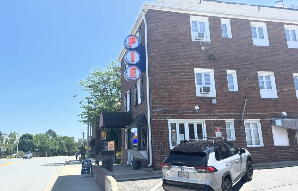 Brick corner building with a vertical neon sign reading 'PIE' outside a storefront on a sunny street. A white SUV is parked in front.
