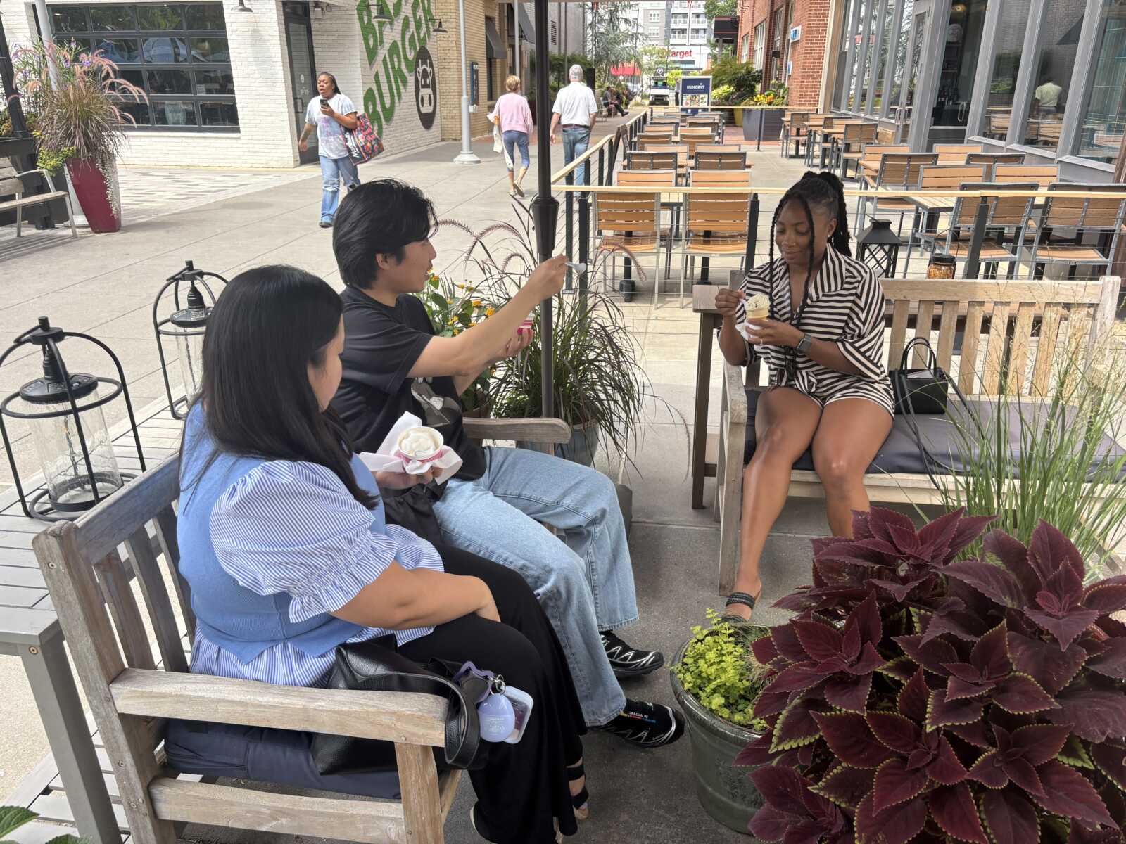 Three friends sit on wooden benches at an outdoor cafe, eating ice cream together; a man hands a pink treat to a woman in jeans while another woman in a striped dress enjoys her cone.
