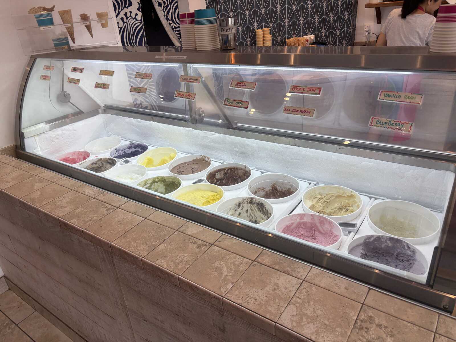 Ice cream parlor display case with colorful gelato flavors in metal tubs behind glass, labeled signs above.